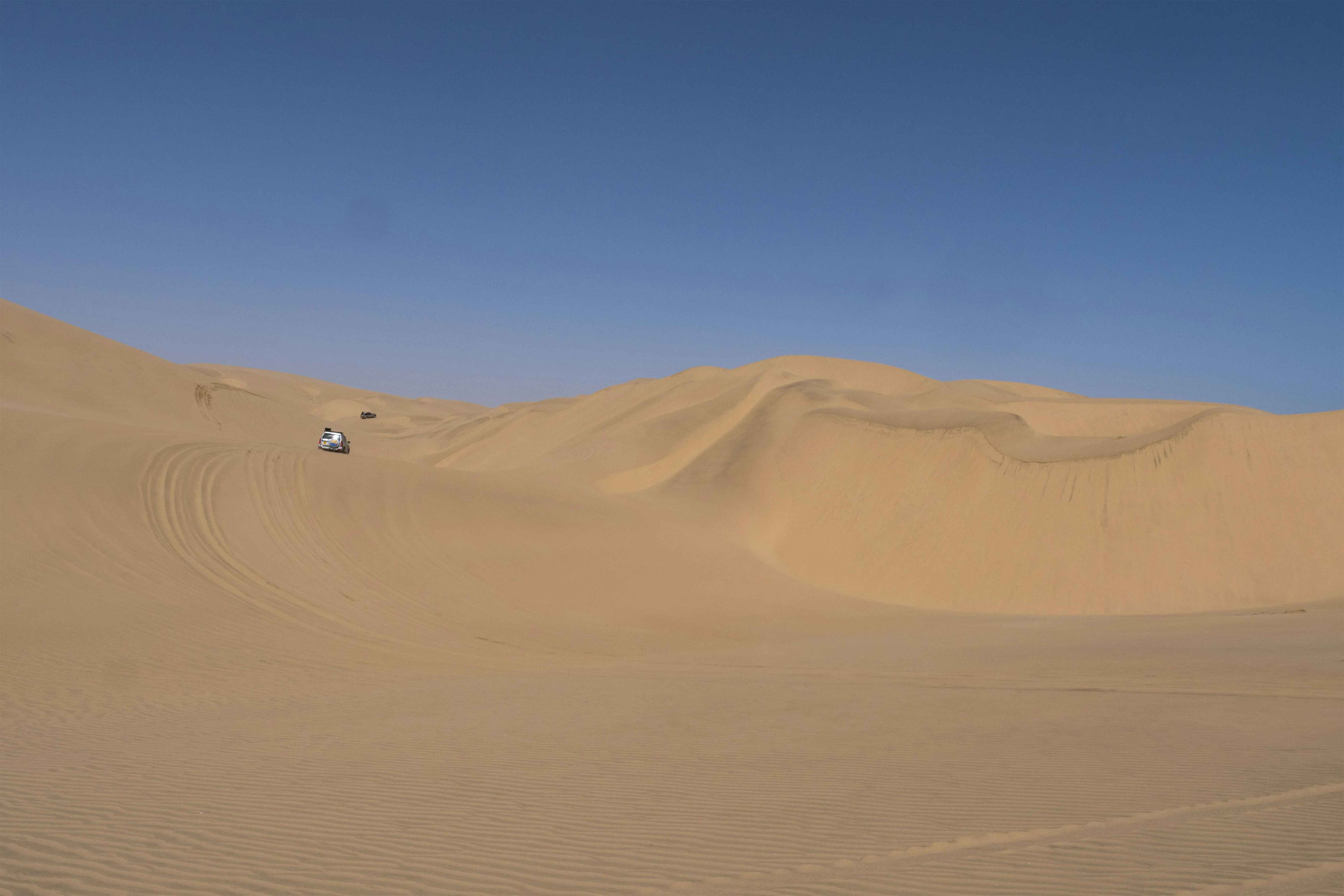 A white SUV driving across vast sand dunes under a clear blue sky
